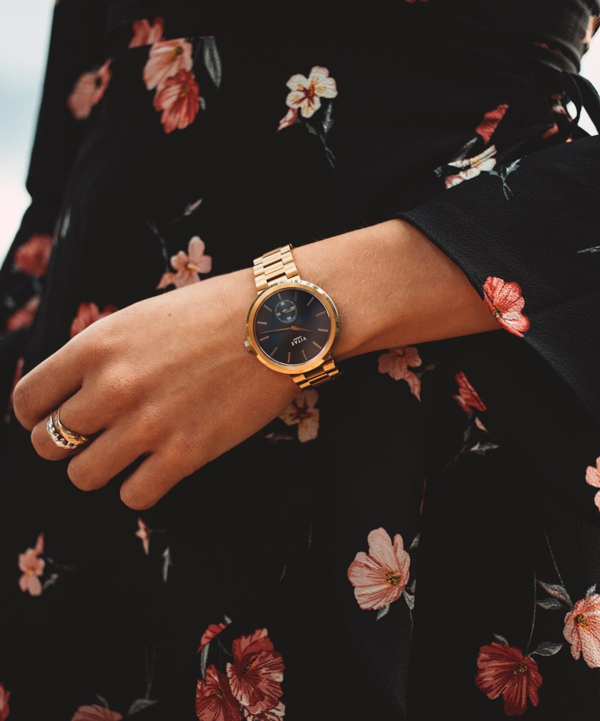 Gold wristwatch worn on a wrist against a dark floral dress