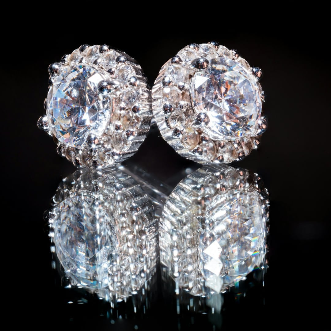 Pair of round diamond halo stud earrings reflected on a black surface