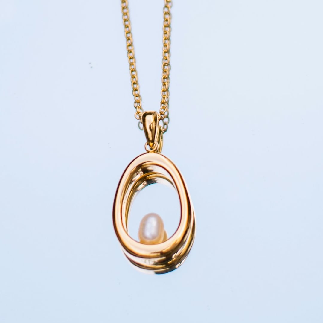 Close-up detail — fine gold necklace with a pearl drop pendant hanging against a pale window