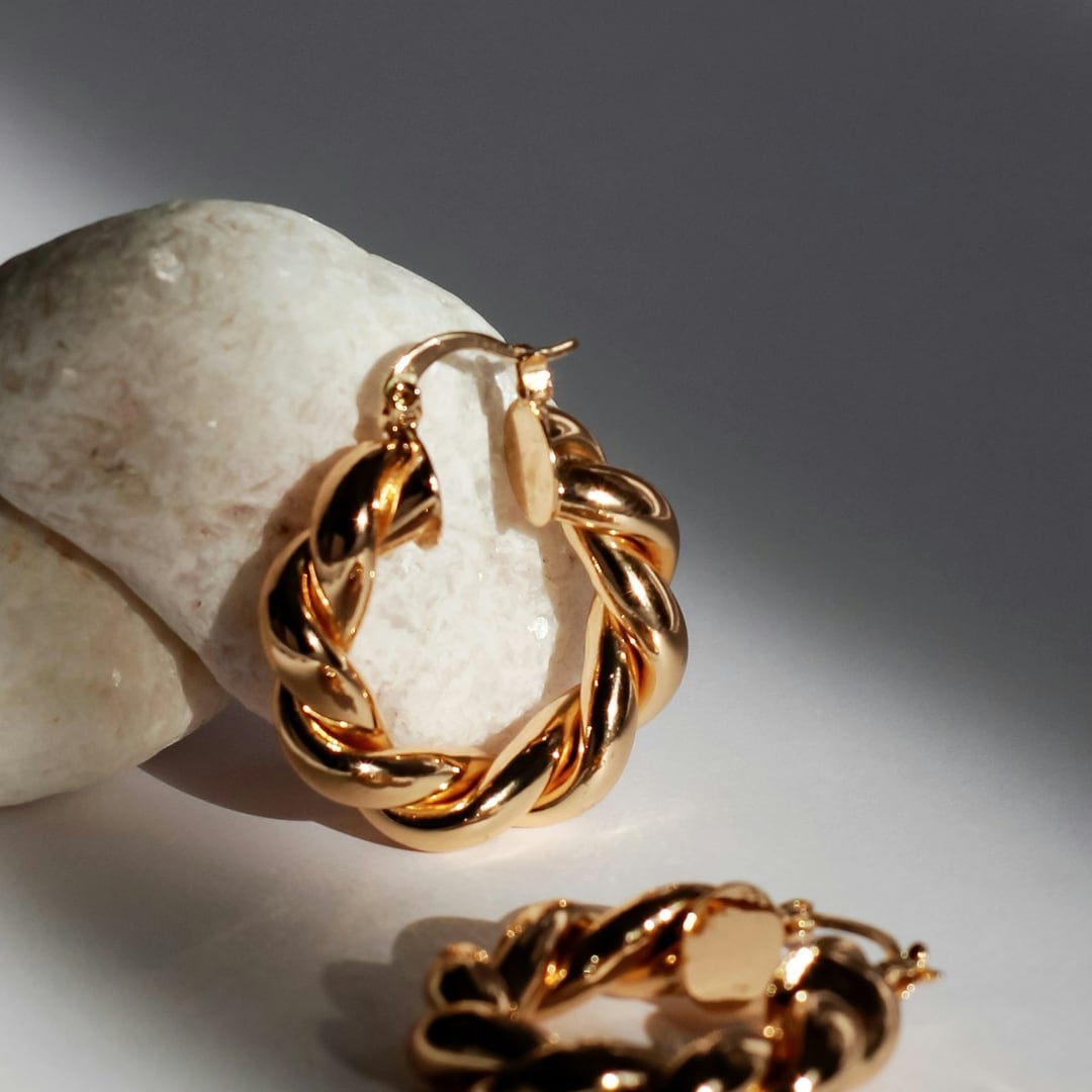 Close-up detail — twisted gold hoop earrings resting on white stones with long shadows